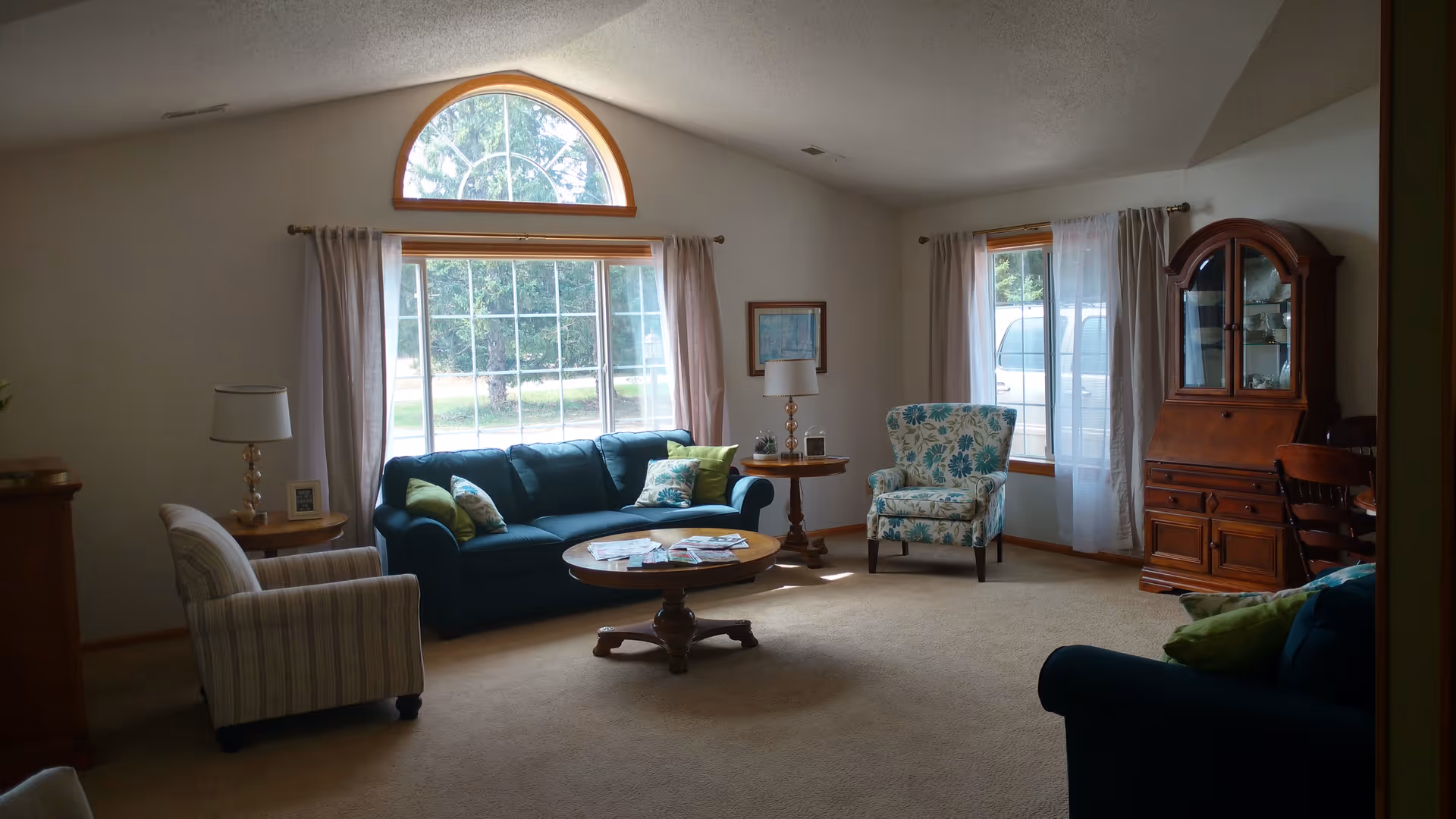 A cozy living room with beige carpet and light-colored walls. The room features a large window with an arched top and curtains, allowing natural light to fill the space. There are two blue sofas with green and patterned cushions, a striped armchair, and a floral-patterned armchair. A round wooden coffee table with magazines is in the center, flanked by two wooden side tables with lamps. A wooden cabinet and a chair are positioned near a smaller window with sheer curtains.