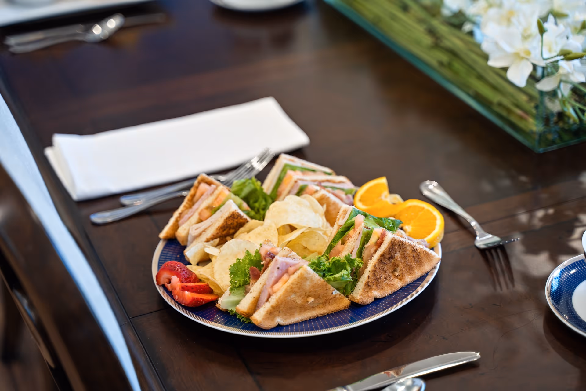 A plate with club sandwiches, potato chips, sliced strawberries, and orange wedges on a dark wooden table set with silverware and a white napkin.