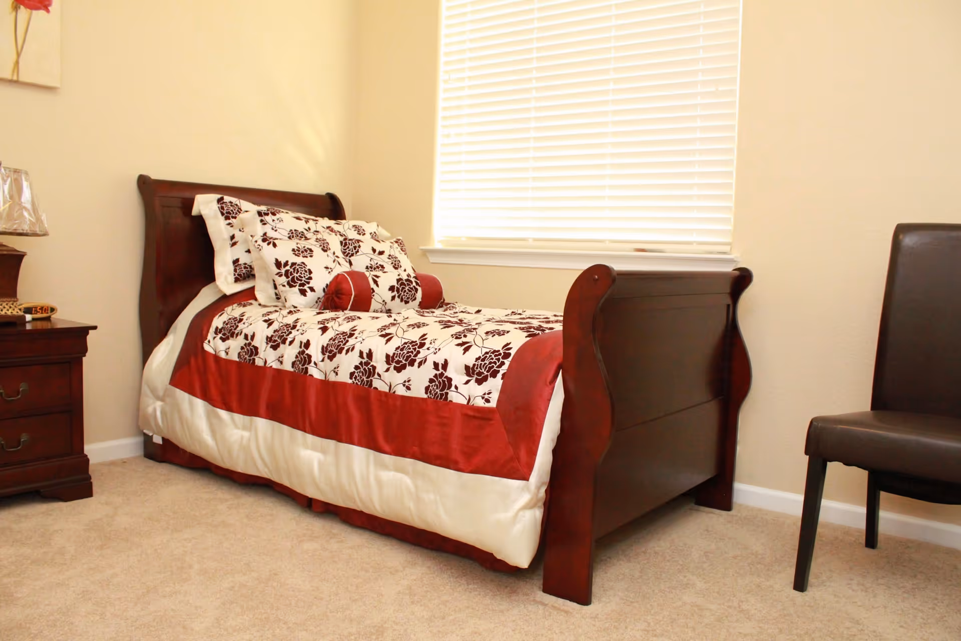 A bedroom with a wooden sleigh bed dressed in white and red floral bedding, a matching pillow and bolster, a wooden nightstand with a lamp and clock, a window with closed blinds, and a dark brown chair.