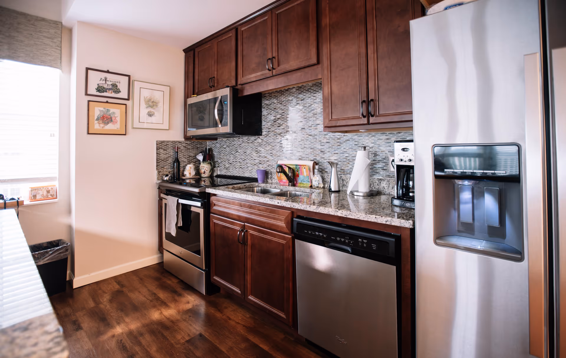 Modern kitchen with stainless steel appliances including a refrigerator, dishwasher, microwave, and oven. The kitchen features dark wooden cabinets, a granite countertop, a mosaic tile backsplash, and framed artwork on the wall near a window with blinds.