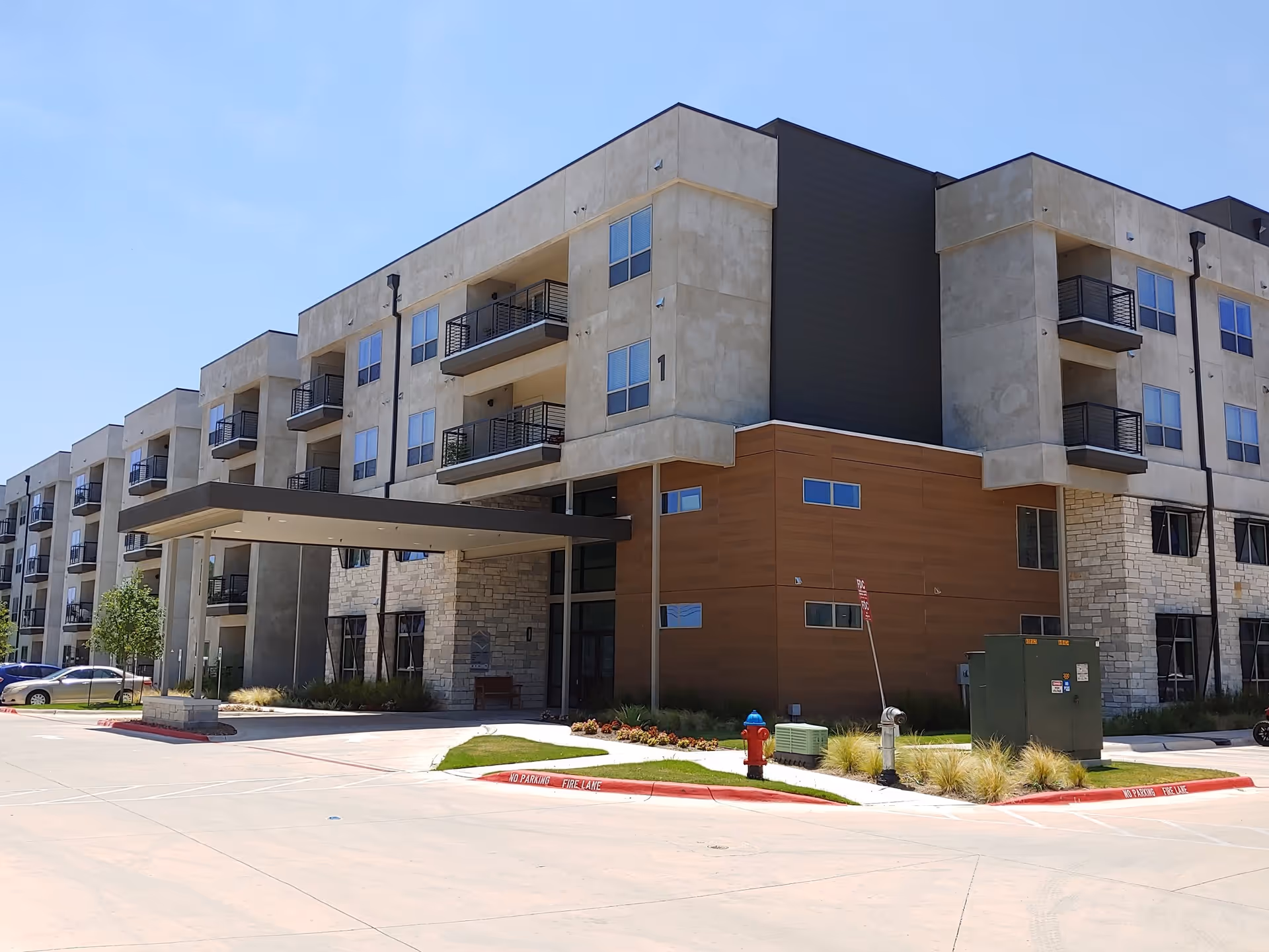 Exterior view of a modern multi-story senior living facility building with balconies, a covered entrance, and a parking area. The building features a combination of stone, wood, and concrete finishes under a clear blue sky.