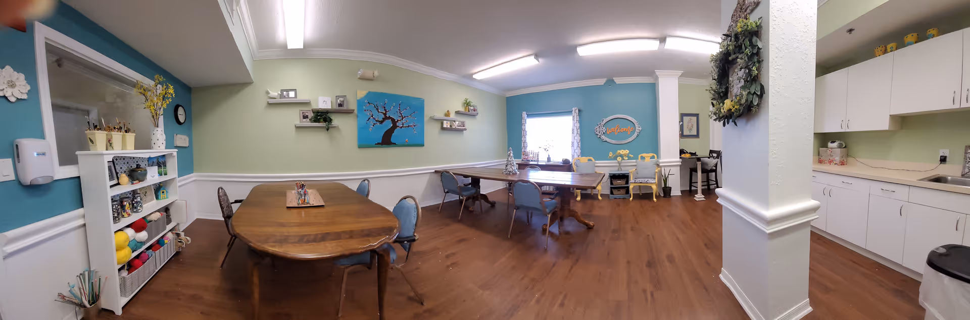 A spacious, well-lit activity room with wooden floors and pastel-colored walls. The room features several tables and chairs arranged for group activities, shelves with craft supplies and yarn, decorative wall art including a tree painting and a welcome sign, and a kitchenette area with white cabinets and a sink. A wreath hangs on a column near the kitchenette.