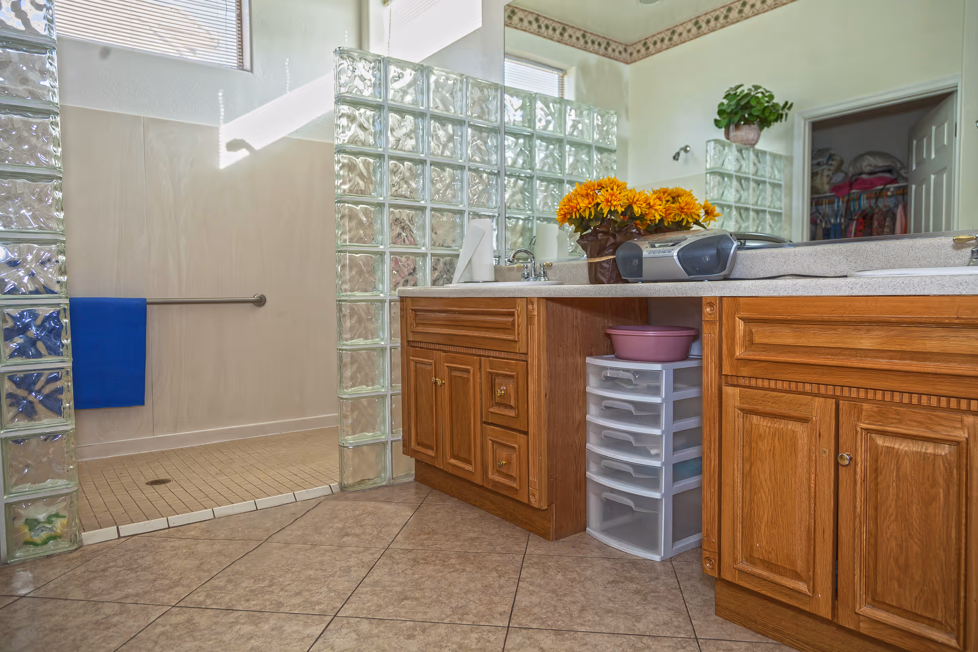 A bathroom with a tiled floor and a walk-in shower enclosed by glass block walls. There is a wooden vanity with a countertop holding a vase of yellow flowers and a small stereo. A plastic drawer unit with a pink basin on top is placed under the counter. A blue towel hangs on a metal bar inside the shower area. A doorway leads to a closet with clothes hanging inside.