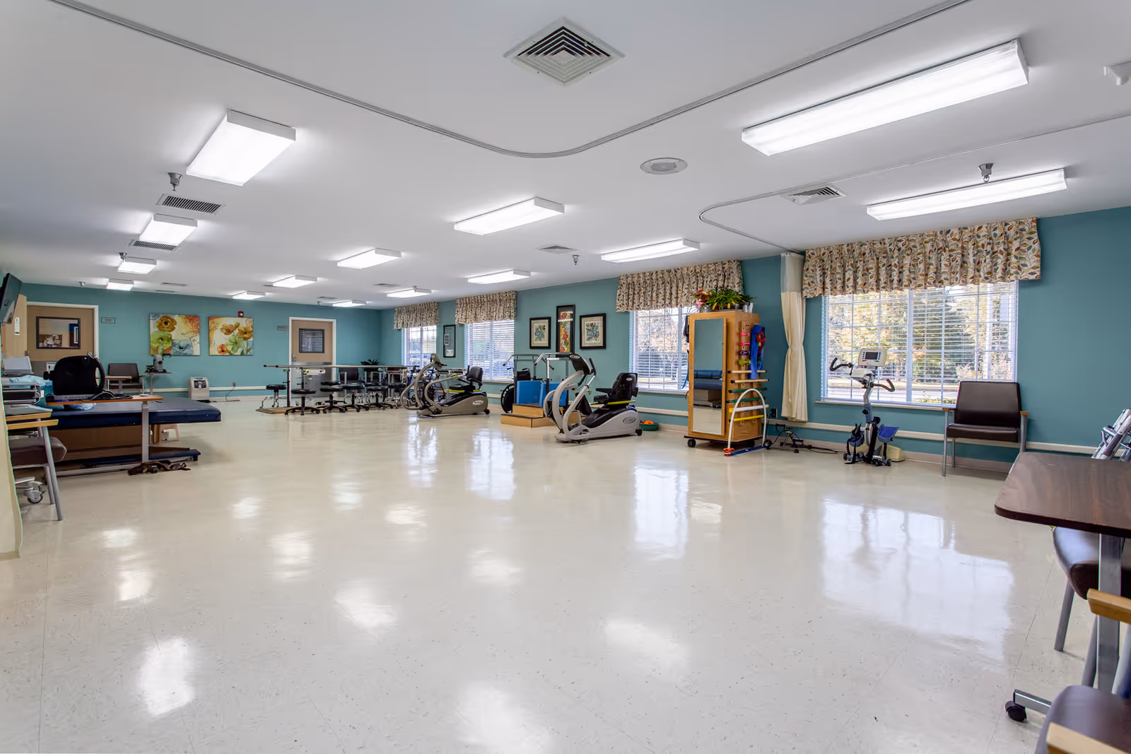 Bright, spacious rehabilitation room with exercise bikes, therapy tables, chairs and large windows.