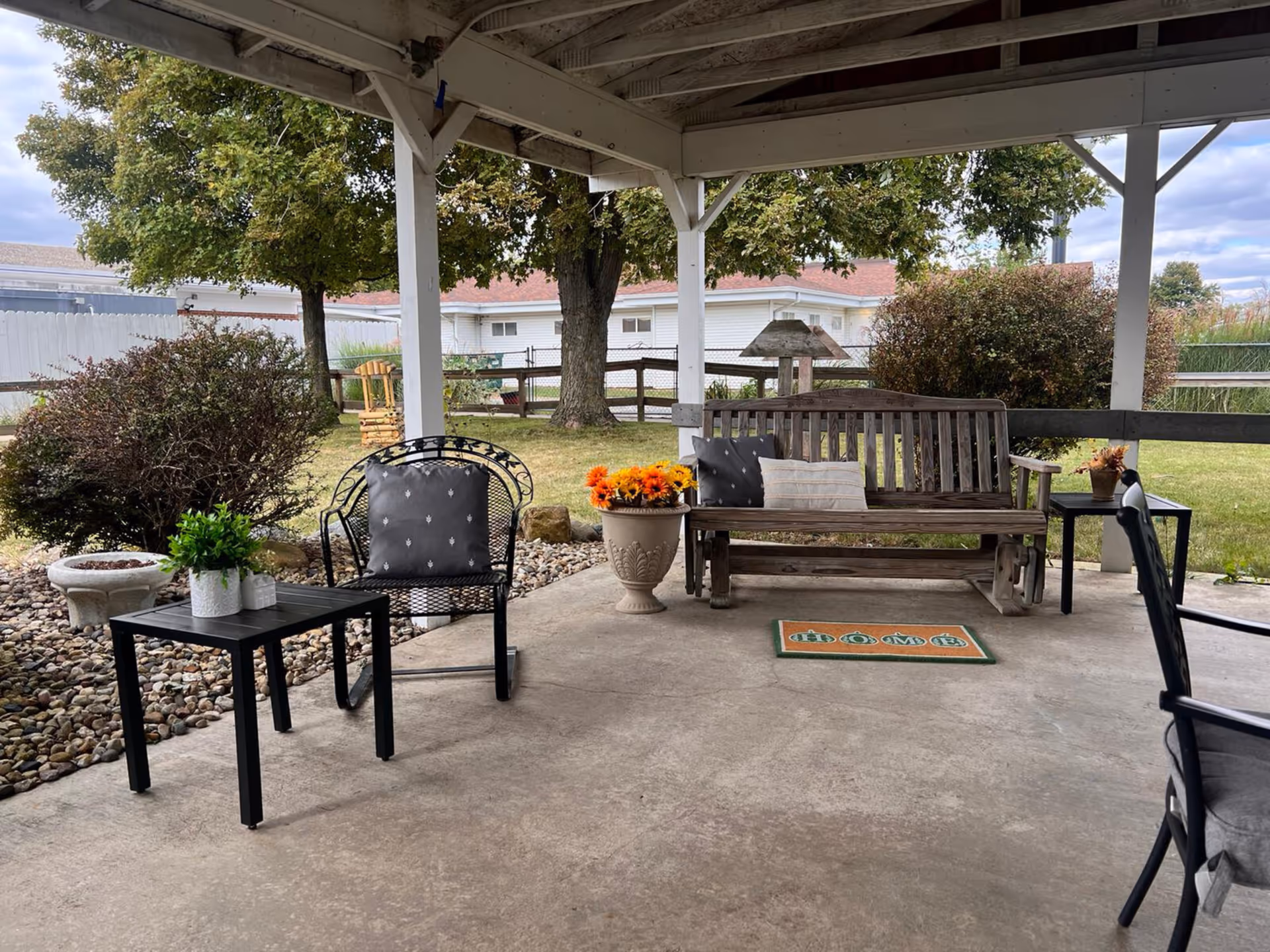 Covered outdoor patio area with a wooden bench adorned with two cushions, a black metal chair with a cushion, two small black tables with potted plants, a large flower pot with orange flowers, and a welcome mat on the concrete floor. The patio overlooks a grassy yard with trees, bushes, and a white fence in the background.