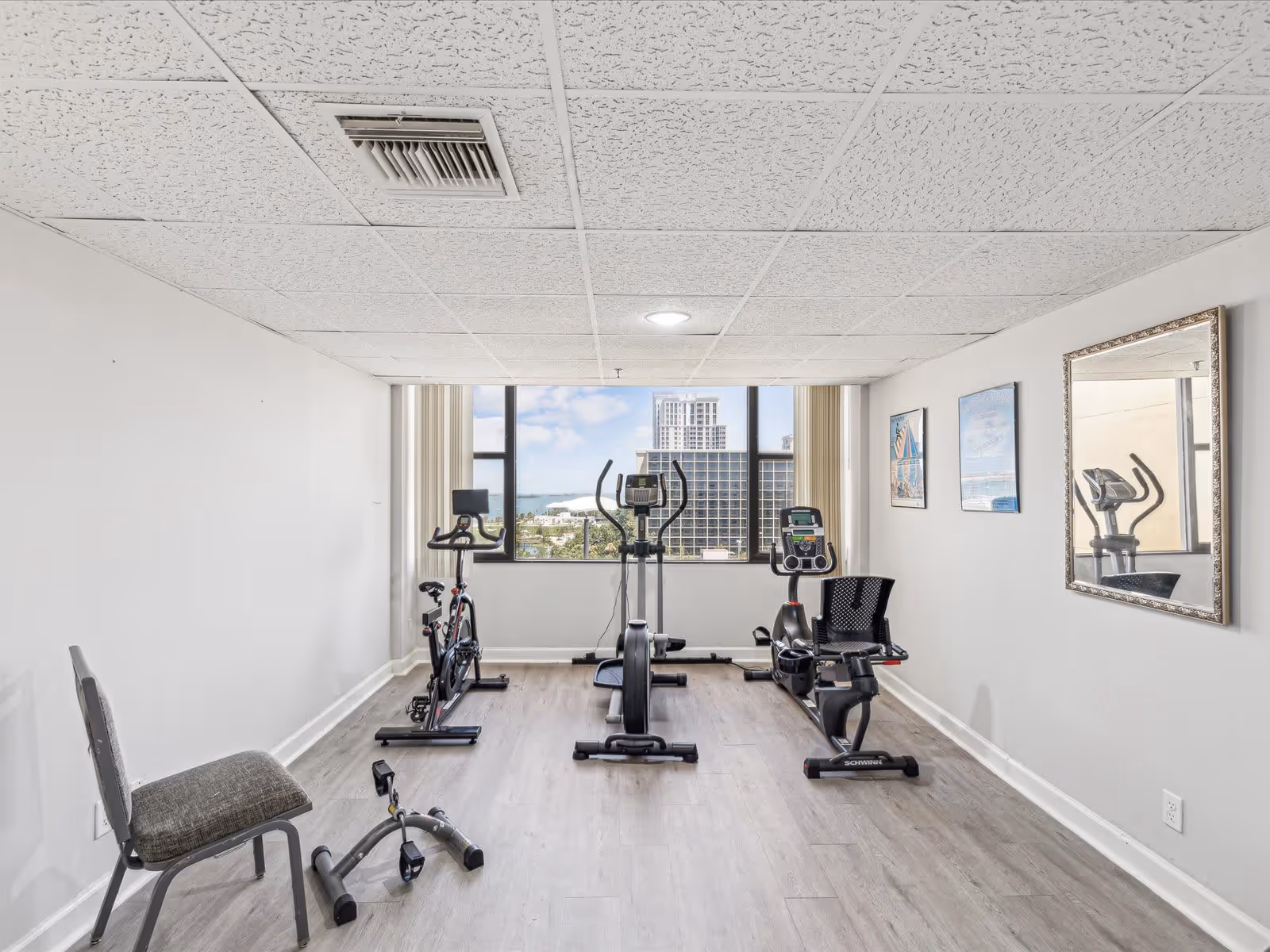 A small exercise room with three stationary bikes lined up facing a large window showing a cityscape and water view. There is a chair and a pedal exerciser on the left side of the room, light wood flooring, white walls, a framed mirror on the right wall, and two framed pictures.