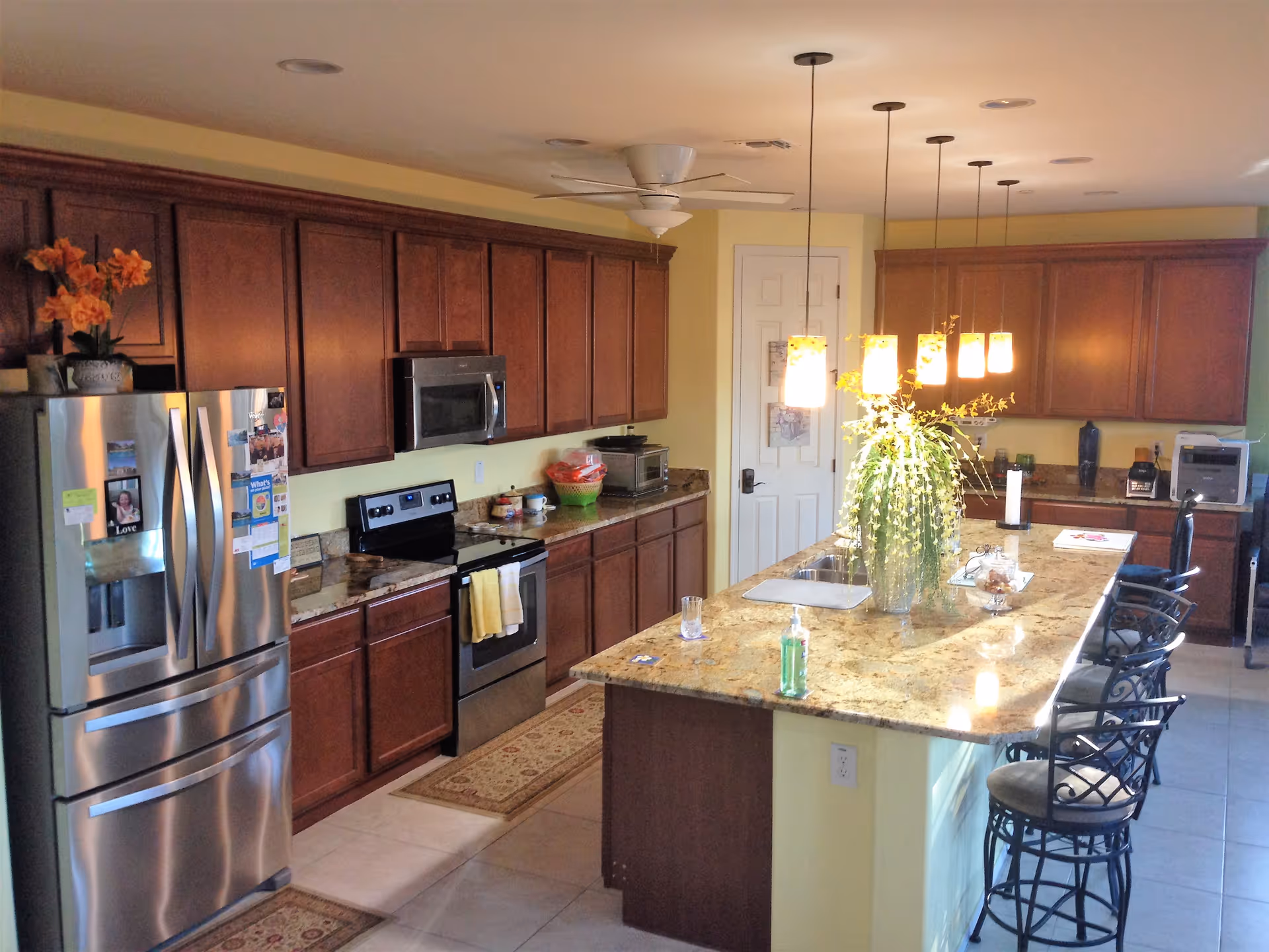 A spacious kitchen with wooden cabinets, a stainless steel refrigerator, stove, and microwave. There is a large granite island in the center with a sink, decorative plant, hand sanitizer, and several bar stools. Pendant lights hang above the island, and the walls are painted yellow.