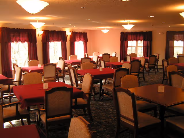 A dining room with multiple tables covered in red tablecloths and surrounded by cushioned chairs. The room has several windows with dark red curtains and is lit by ceiling lights.