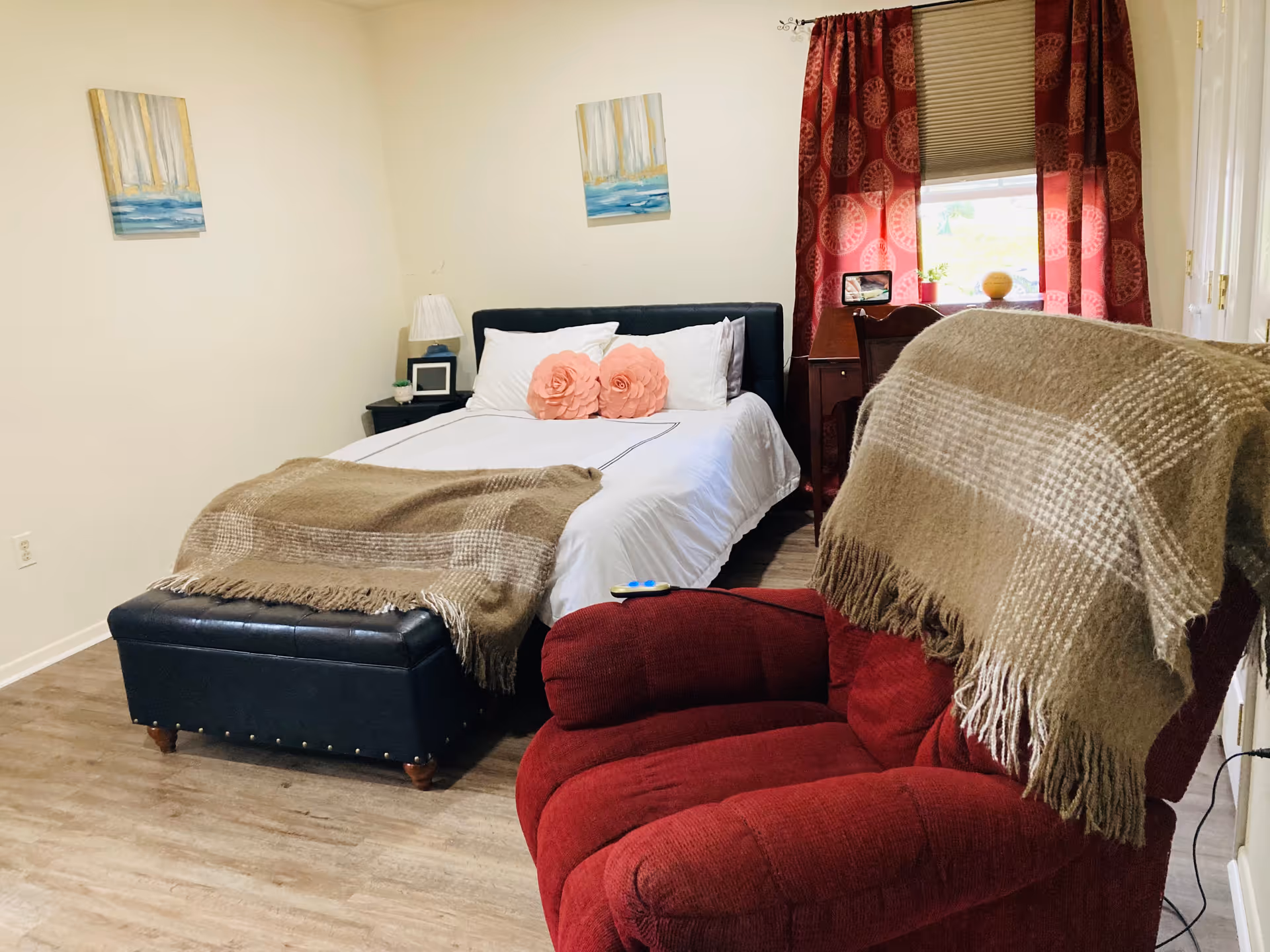 A cozy bedroom with a neatly made bed featuring white linens and two pink flower-shaped pillows. At the foot of the bed is a dark blue ottoman with a brown plaid blanket draped over it. A red upholstered armchair with a similar brown plaid blanket is positioned in the foreground. The room has light-colored walls with two paintings above the bed, a small bedside table with a lamp, and a window with red patterned curtains letting in natural light.