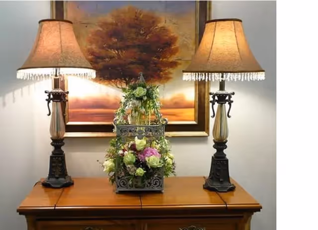 A wooden table with two ornate lamps on either side and a decorative floral arrangement in the center. Behind the table is a framed painting of a large tree with a sunset or sunrise background.