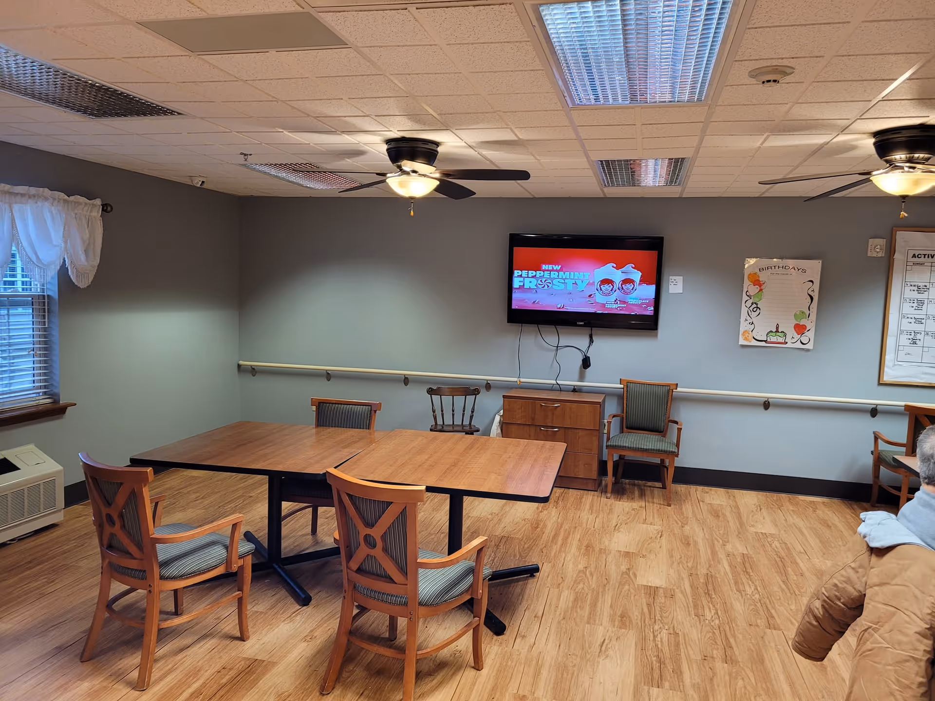 A senior living common room with wooden tables and chairs, a wall-mounted TV, ceiling fans, and bulletin boards.