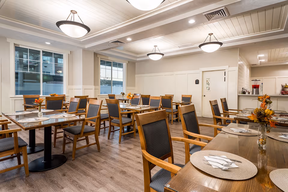 A bright and clean dining room in a senior living facility with multiple wooden tables and chairs arranged neatly. Each table is set with placemats, napkins, and silverware, and small vases with autumn-themed floral arrangements are placed on the tables. The room has large windows allowing natural light to enter, light-colored walls, and ceiling lights providing additional illumination. A beverage station with dispensers is visible in the background.