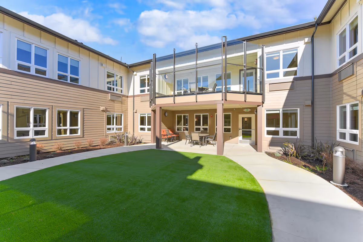 Outdoor courtyard area of Silverado Bellevue Memory Care Community with green artificial turf, concrete pathways, and a two-story building surrounding the space. The upper level has a balcony with glass railings and outdoor seating, while the lower level features a covered patio with tables and chairs. The sky is partly cloudy.