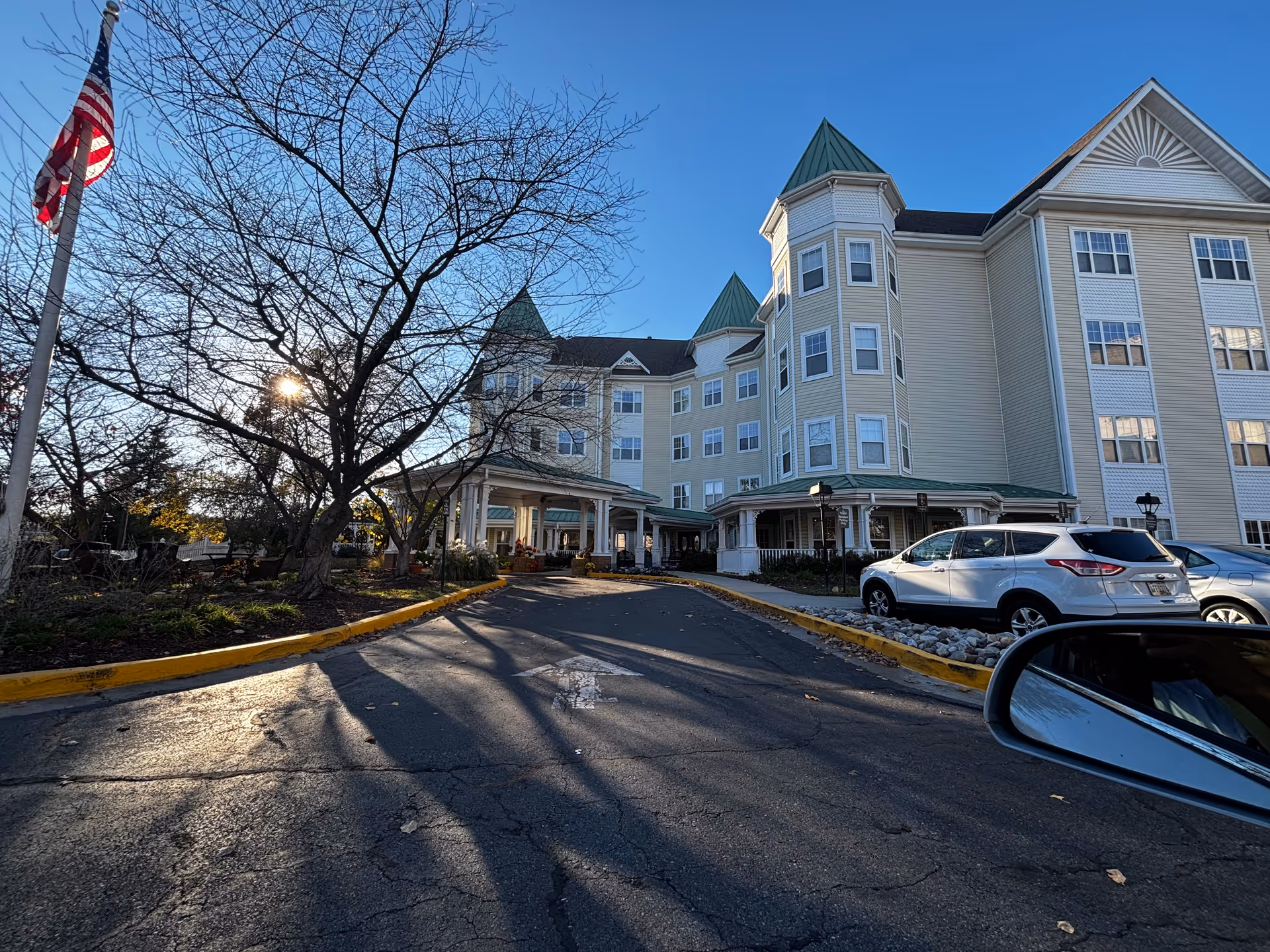Front entrance of a multi-story senior living building with a covered porte-cochere, parked cars, leafless trees, and an American flag.