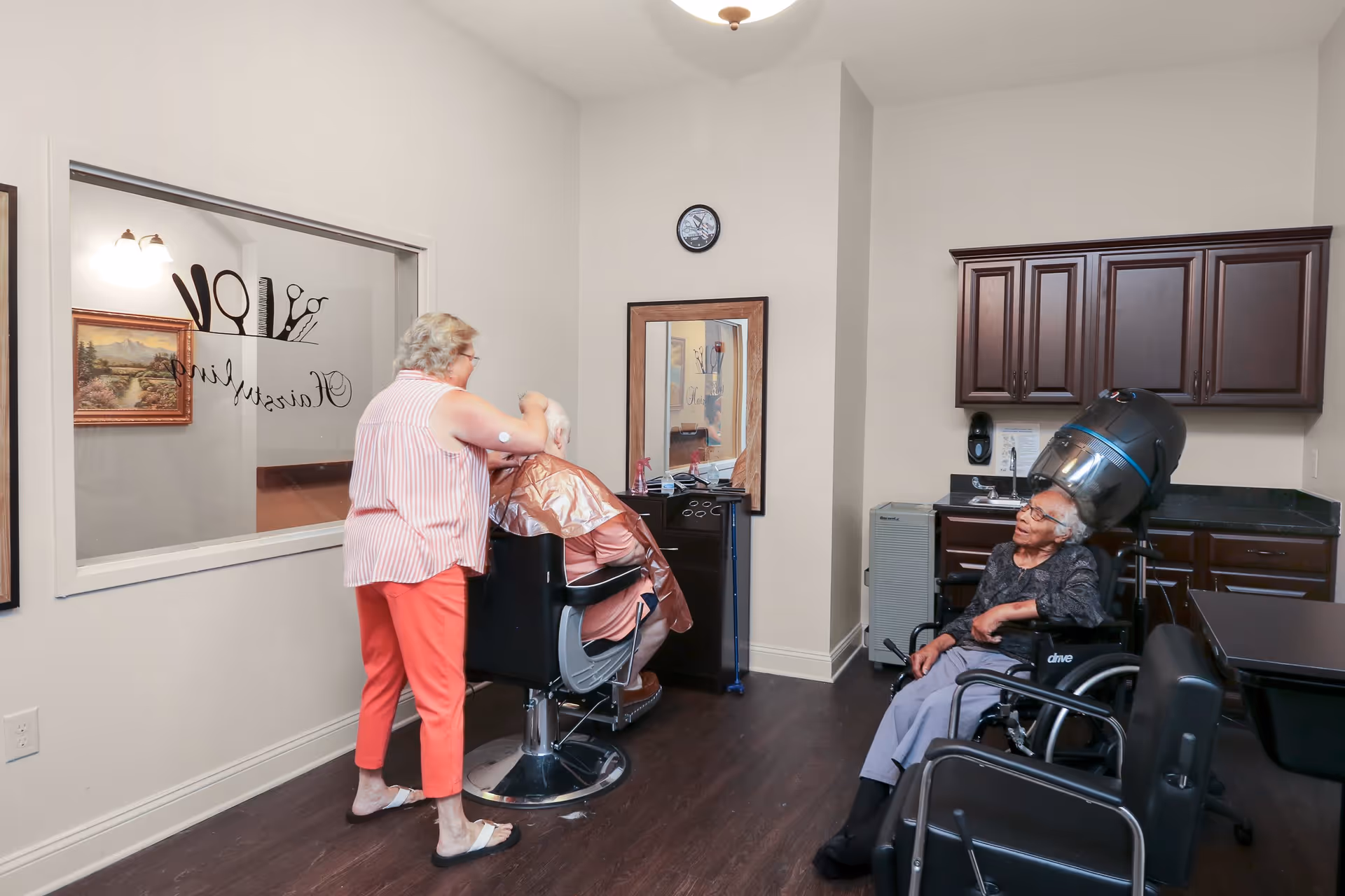 A senior living facility hair salon with a woman cutting an elderly man's hair seated in a barber chair. Another elderly woman is sitting in a wheelchair under a hair dryer. The room has wooden cabinets, a mirror, and a clock on the wall.