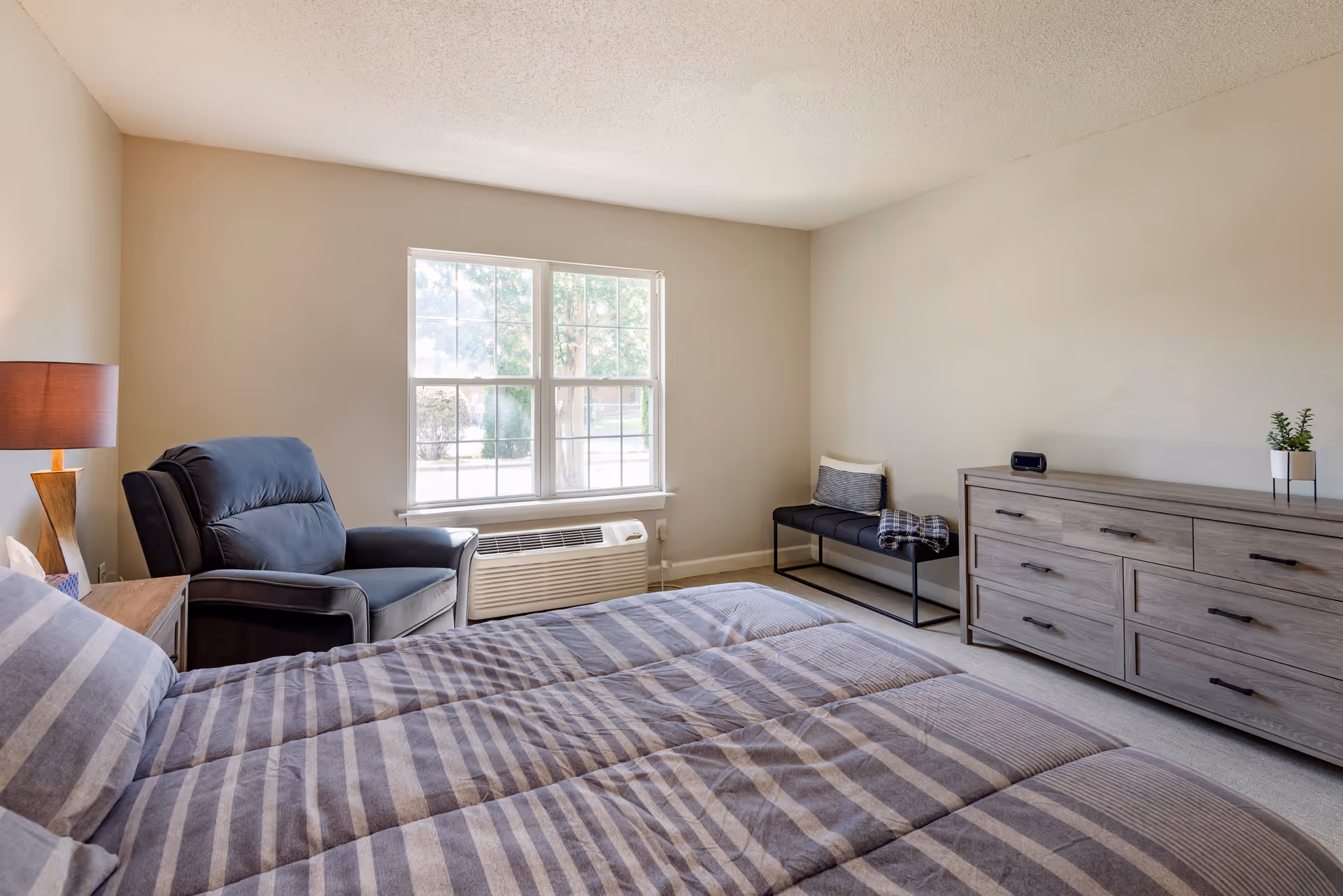 A bright bedroom with a large window letting in natural light. The room features a bed with striped bedding, a dark armchair next to a wooden nightstand with a lamp, a black bench with a pillow and blanket, and a wooden dresser with a small plant and a clock on top.