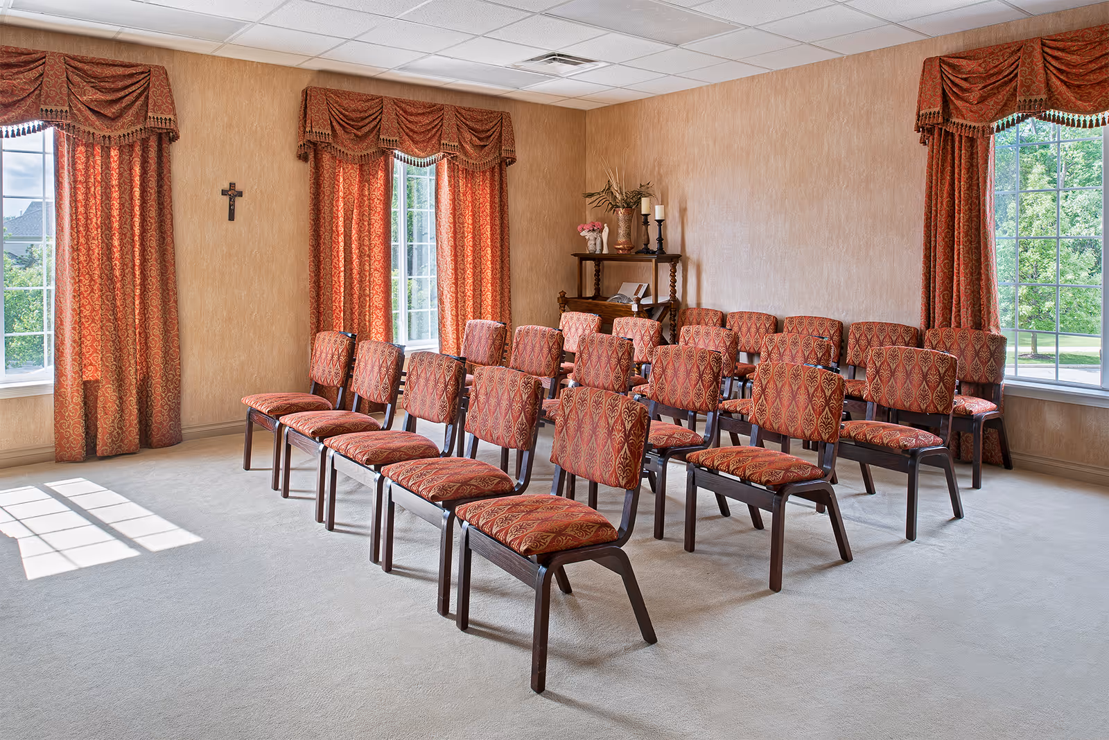 A small meeting or chapel room with rows of patterned red chairs arranged facing forward. The room has beige walls, large windows with red patterned curtains, and a small wooden shelf with decorative items including candles and flowers. A cross is mounted on one wall.