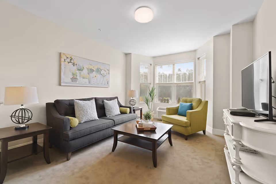 A bright living room with a dark gray sofa adorned with white and green pillows, a green armchair with a blue pillow, a wooden coffee table with decorative items, two side tables with lamps, a large window with white blinds, and a white TV stand with a flat-screen television.