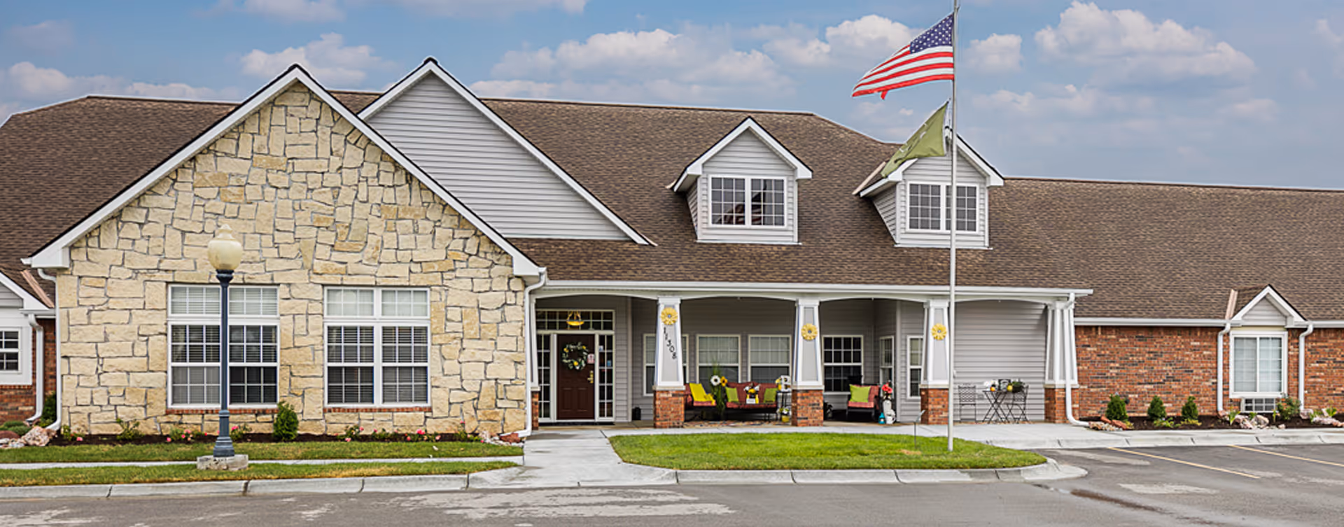 Front exterior of a single-story senior living facility with stone and brick facade, a covered porch with seating, and an American flag.