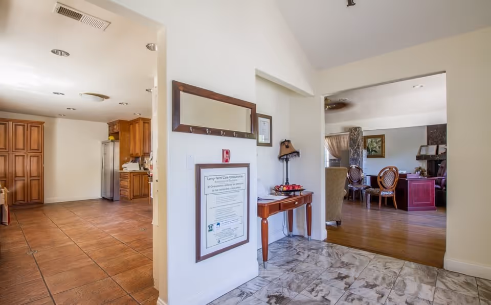 Interior view of a senior living facility showing a hallway with marble flooring leading to a living room area with wooden flooring and classic furniture. To the left, there is a kitchen area with wooden cabinets and tiled flooring. A small wooden table with a lamp and decorative items is placed against the wall, which also has a framed notice and a mirror.