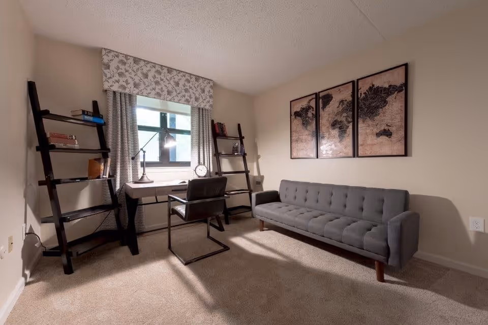A cozy interior room featuring a gray tufted sofa against a beige wall with three framed world map artworks above it. To the left, there is a black desk with a modern lamp, a clock, and a chair in front of a window with patterned curtains and a valance. Two black ladder-style bookshelves flank the desk, holding books and decorative items. The room has beige carpeting and neutral-colored walls.