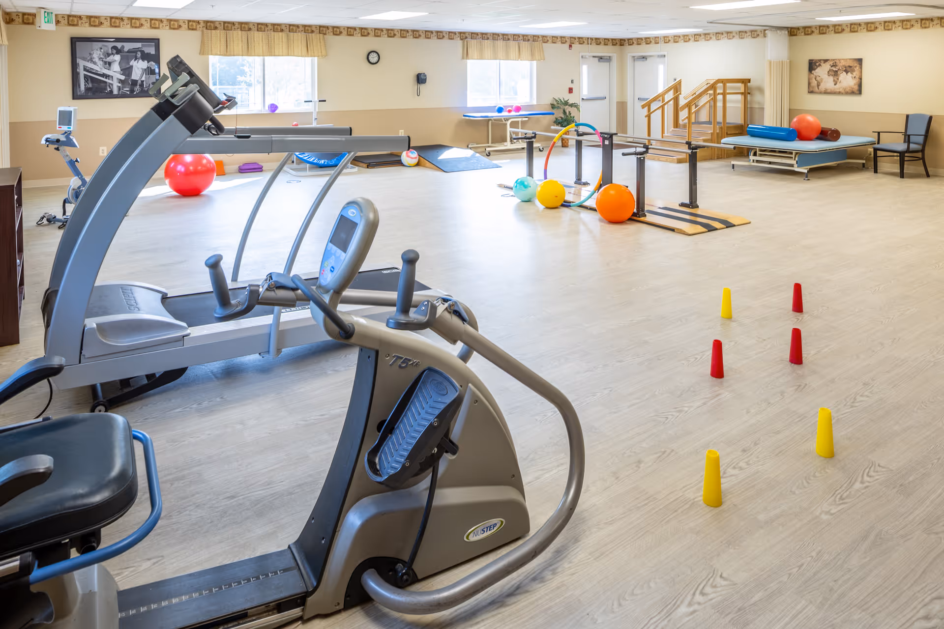 A spacious exercise and rehabilitation room with various fitness equipment including a treadmill, stationary bike, exercise balls, parallel bars, and colorful cones arranged on the floor. The room has large windows with yellow valances, light wood flooring, and light-colored walls with framed pictures. There is also a small set of stairs with a handrail and a therapy table in the background.