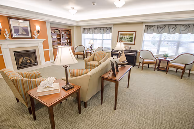 A cozy living room area with beige and striped armchairs, a beige sofa, wooden side tables with lamps, a fireplace with decorative items above it, a bookshelf, and large windows with valances letting in natural light.