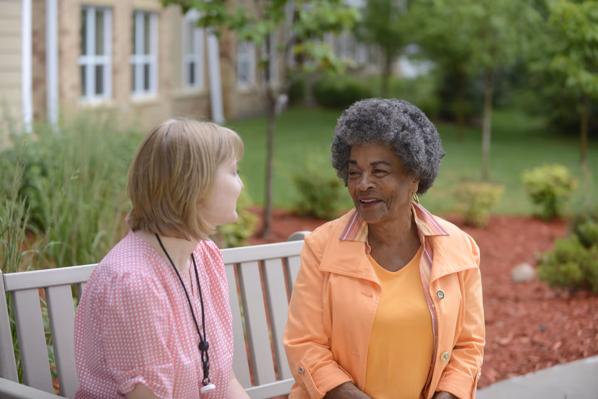 Two women sitting on a bench outside in a garden area, engaged in conversation. One woman has short blonde hair and is wearing a pink patterned blouse, while the other woman has curly gray hair and is wearing an orange jacket over a yellow top. There are green plants, trees, and a building with windows in the background.