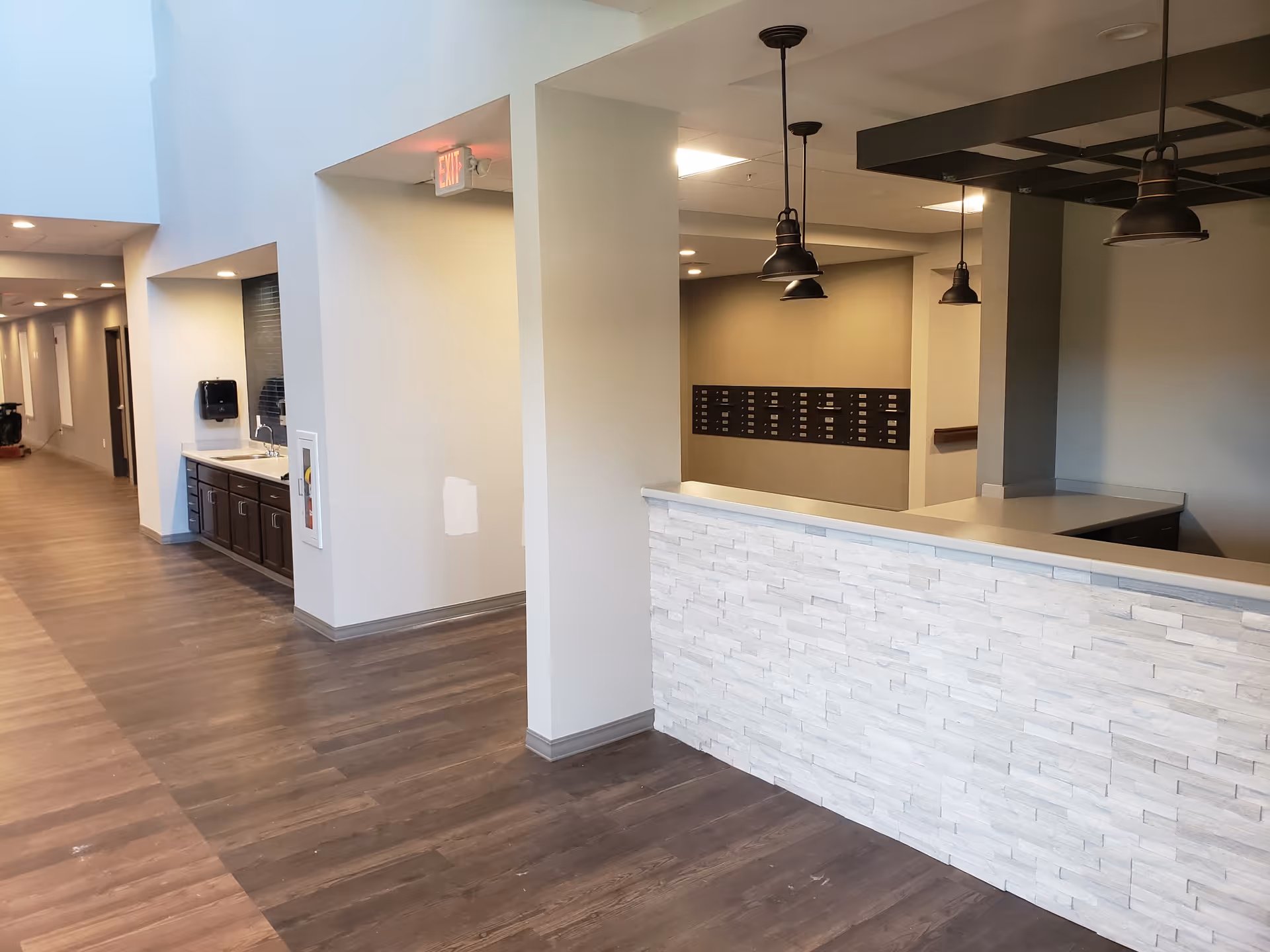 Interior view of a senior living facility hallway with wood flooring, a white stone reception counter with pendant lights above, a sink area with cabinets along one wall, and mailboxes mounted on the far wall.