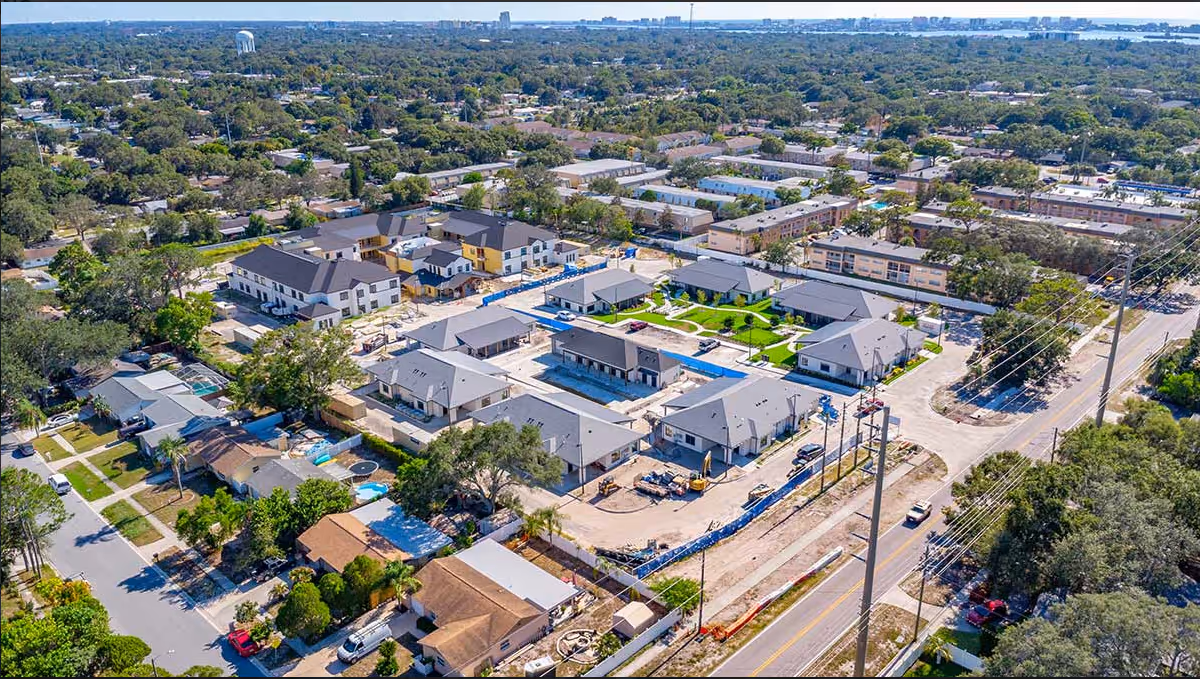 Aerial view of Elevate Senior Living at Clearwater facility showing multiple residential buildings with gray roofs arranged around green lawns and pathways. Surrounding the facility are residential neighborhoods with trees and streets, and a main road runs alongside the property.