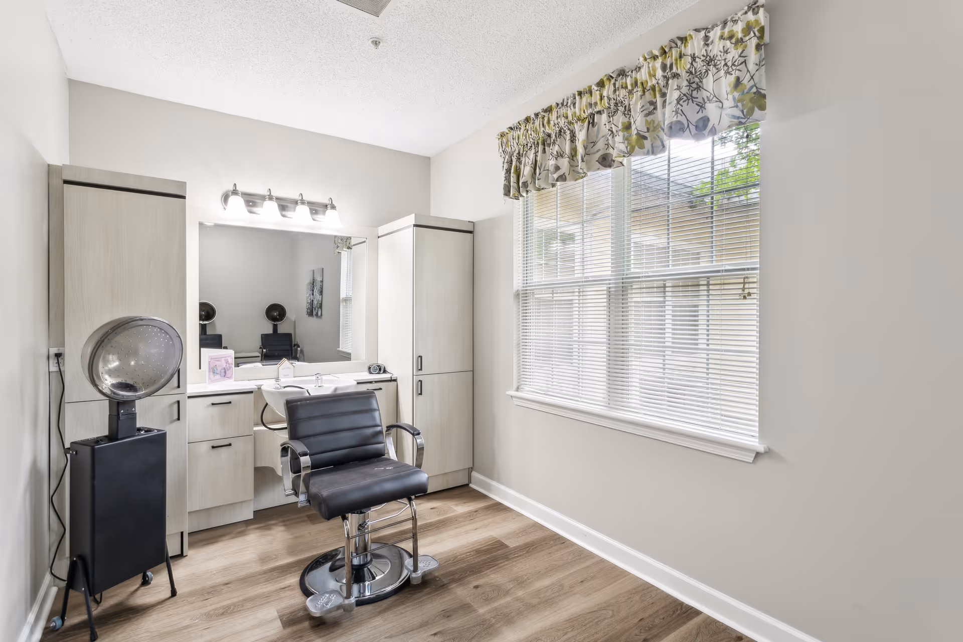 Small salon/hair-styling room with a black styling chair, hooded dryer, vanity sink and storage cabinets beside a large window.