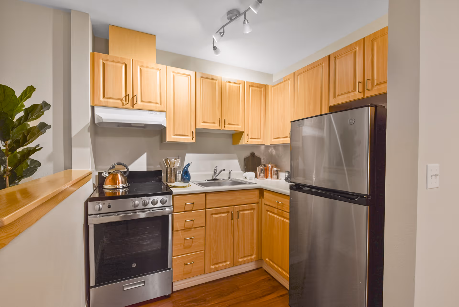 A compact kitchen with light wood cabinets, a stainless steel refrigerator, a stainless steel oven with a kettle on top, a white countertop with a sink, and various kitchen utensils and containers. There is a small plant visible on the left side and track lighting on the ceiling.