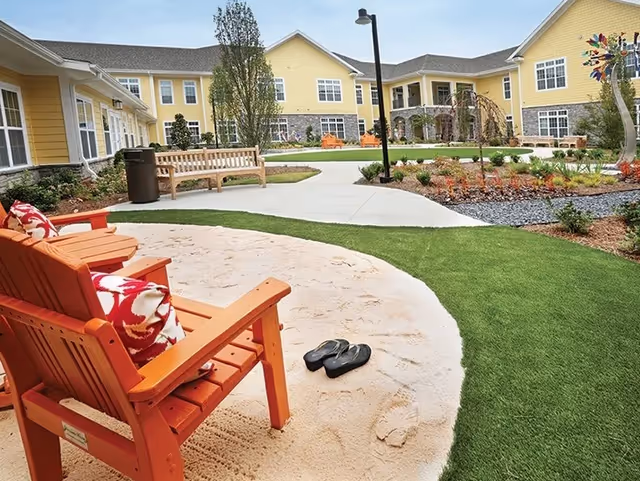 Outdoor courtyard area at Cobblestone At Park Springs featuring orange wooden chairs with patterned cushions on a sand patch, a pair of black flip-flops on the sand, green artificial grass, a wooden bench, landscaped garden beds, and a yellow two-story building in the background under a clear sky.