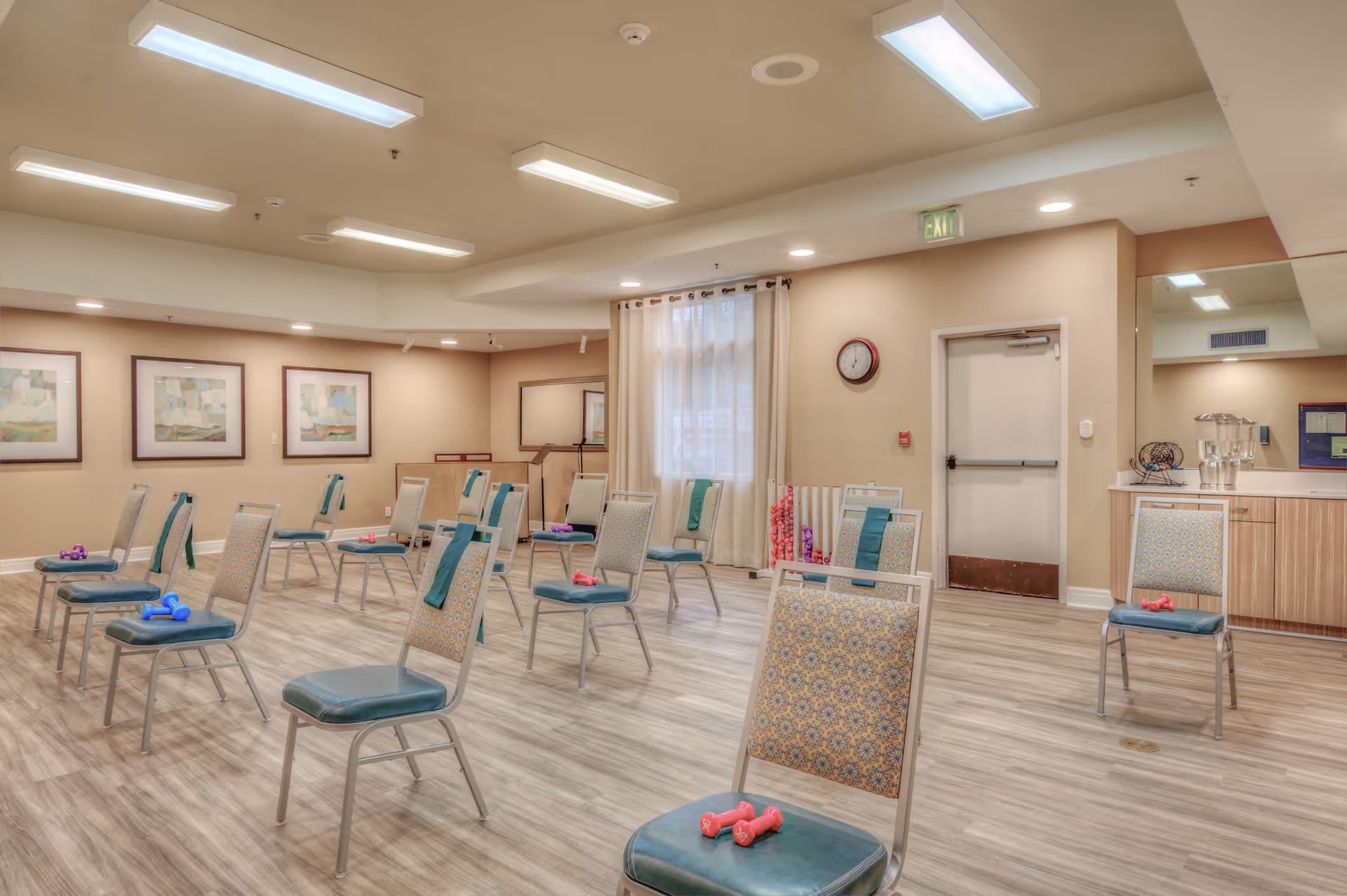 A spacious exercise room with multiple chairs arranged in rows, each chair holding a pair of small dumbbells and a towel. The room has light-colored walls, wood-patterned flooring, framed artwork on the walls, a clock, and a water dispenser on a counter. There is a window with sheer curtains and an exit door visible.
