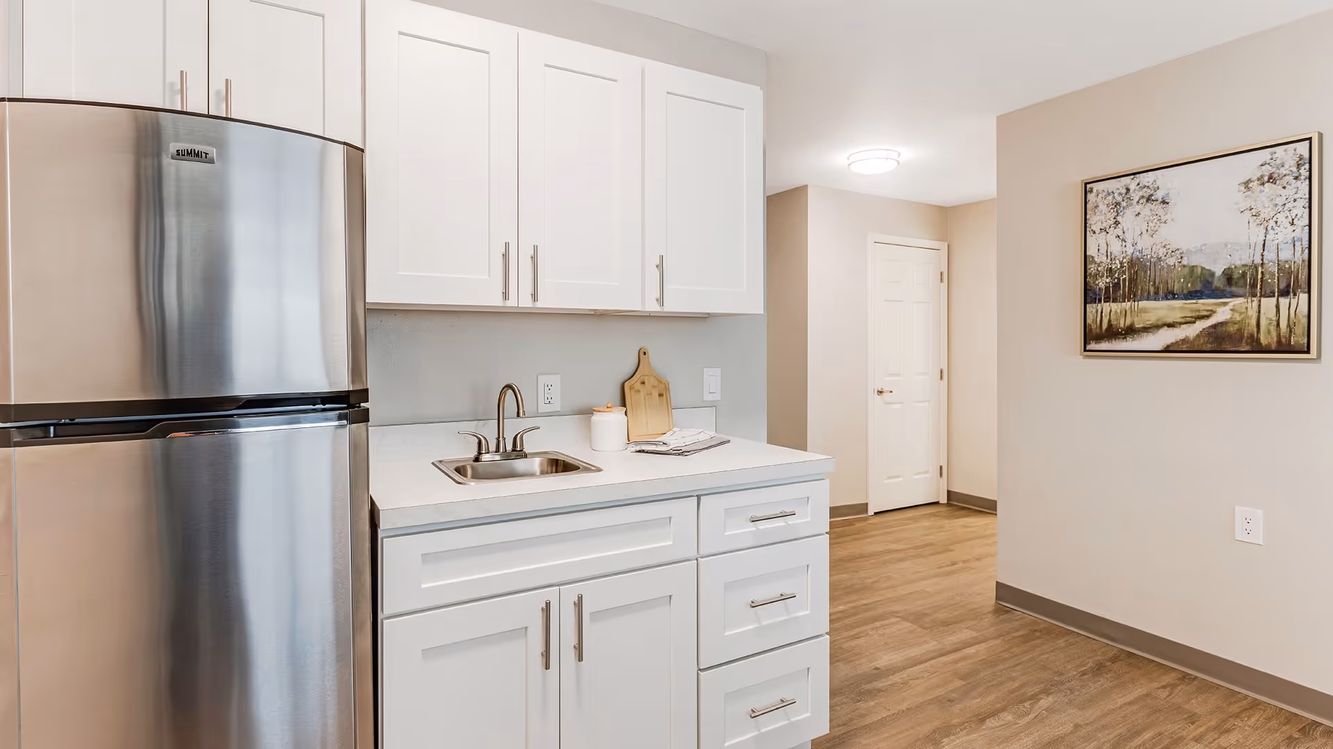 A modern kitchen area with white cabinets, a small stainless steel sink, and a stainless steel refrigerator. The kitchen has a light-colored countertop and wood flooring. In the background, there is a hallway with a closed white door and a framed landscape painting on the beige wall.