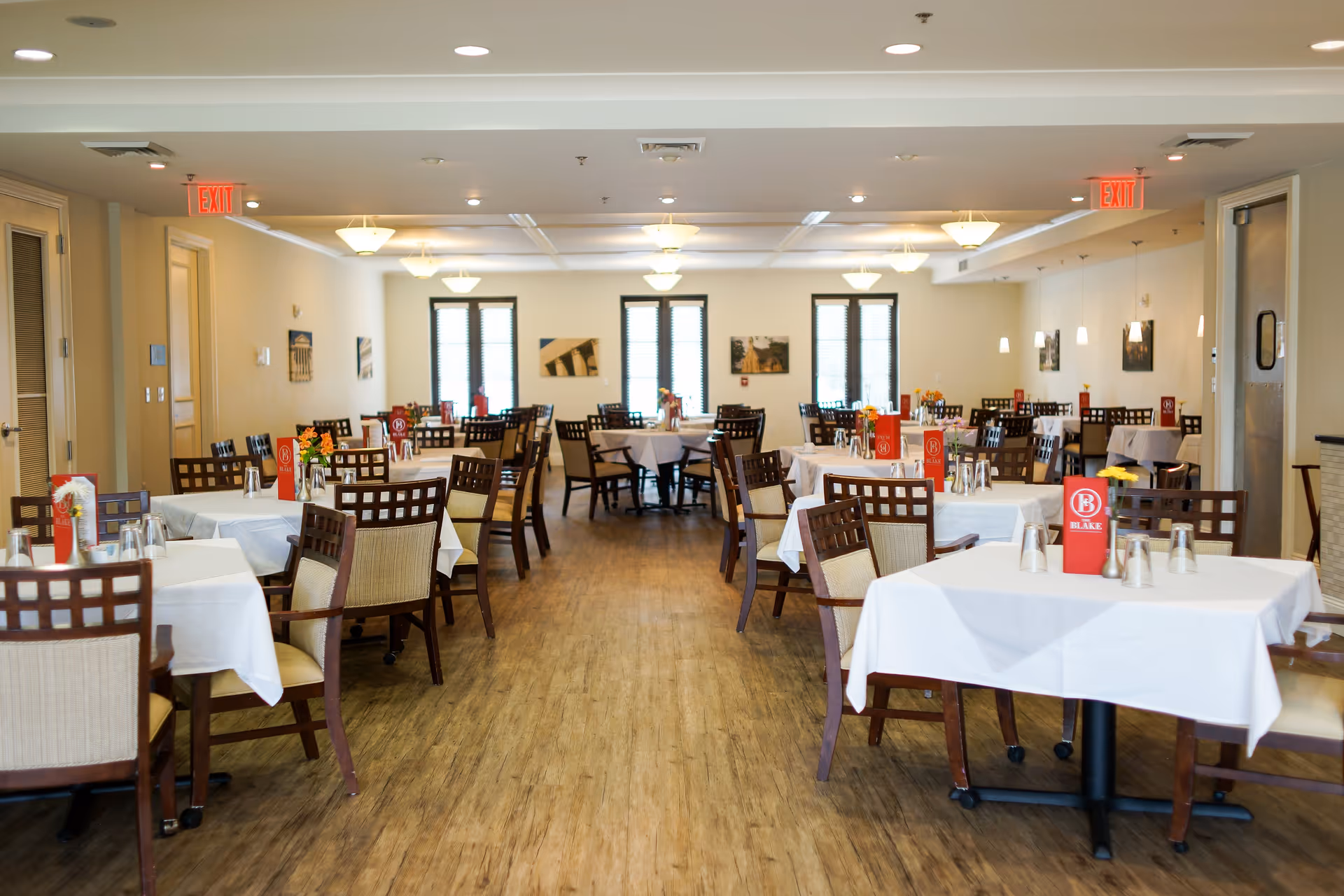 Spacious dining room with neatly set tables, white tablecloths, wooden chairs and menus on each table.