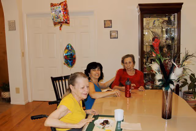 Three older women seated around a dining table with balloons, a vase of flowers, and a china cabinet in the background.