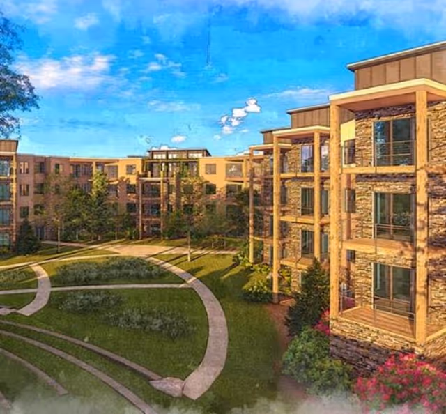 Exterior view of a multi-story senior living facility with stone and wood accents, surrounded by landscaped green lawns and curved walking paths under a blue sky with some clouds.