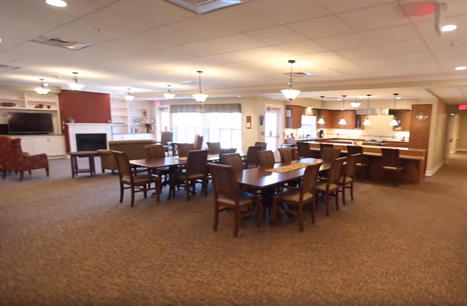 A spacious common area in a senior living facility featuring a dining area with multiple wooden tables and chairs, a kitchen with a large island and pendant lighting, and a living room area with armchairs, a sofa, a fireplace, and a television. The room is well-lit with ceiling lights and natural light from large windows.