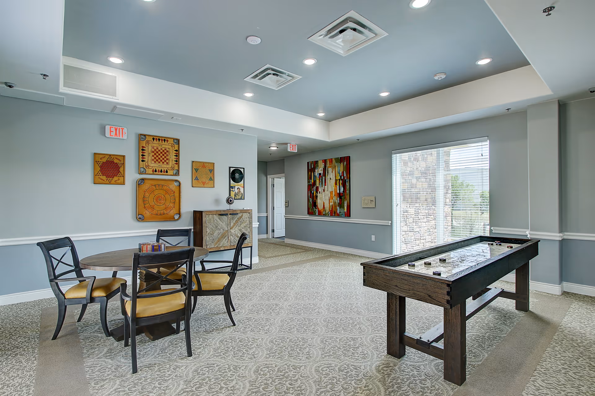 Well-lit senior living common room with a round table and chairs, wall art, and a shuffleboard table by a large window.
