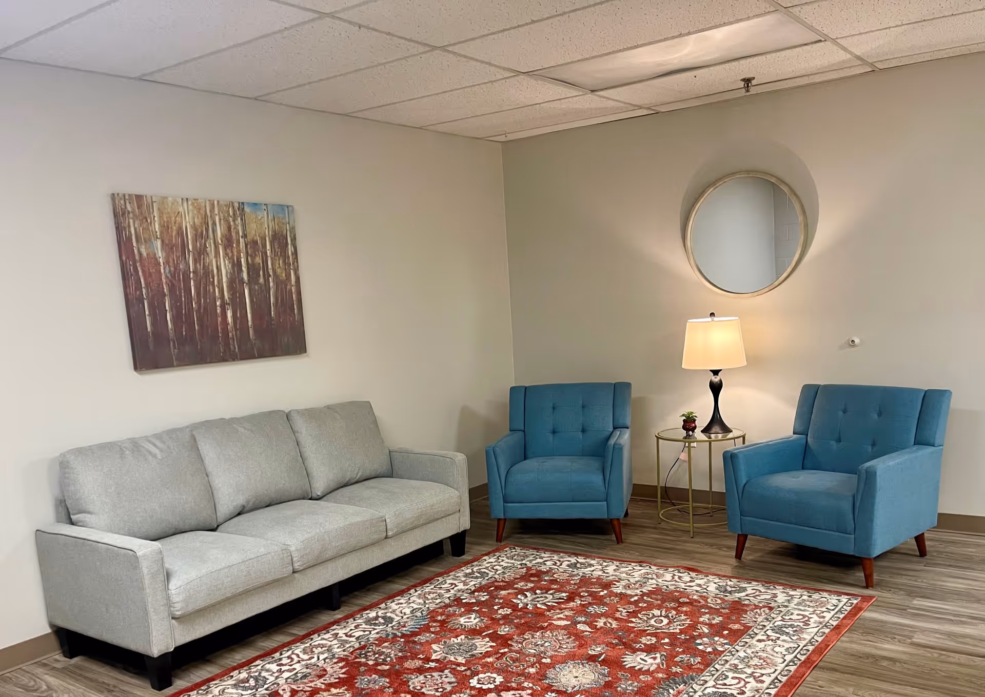 A cozy sitting area in a senior living facility featuring a light gray three-seater sofa, two blue armchairs, a round side table with a lamp and small plant, a round wall mirror, a painting of trees on the wall, and a red patterned area rug on a wood floor.