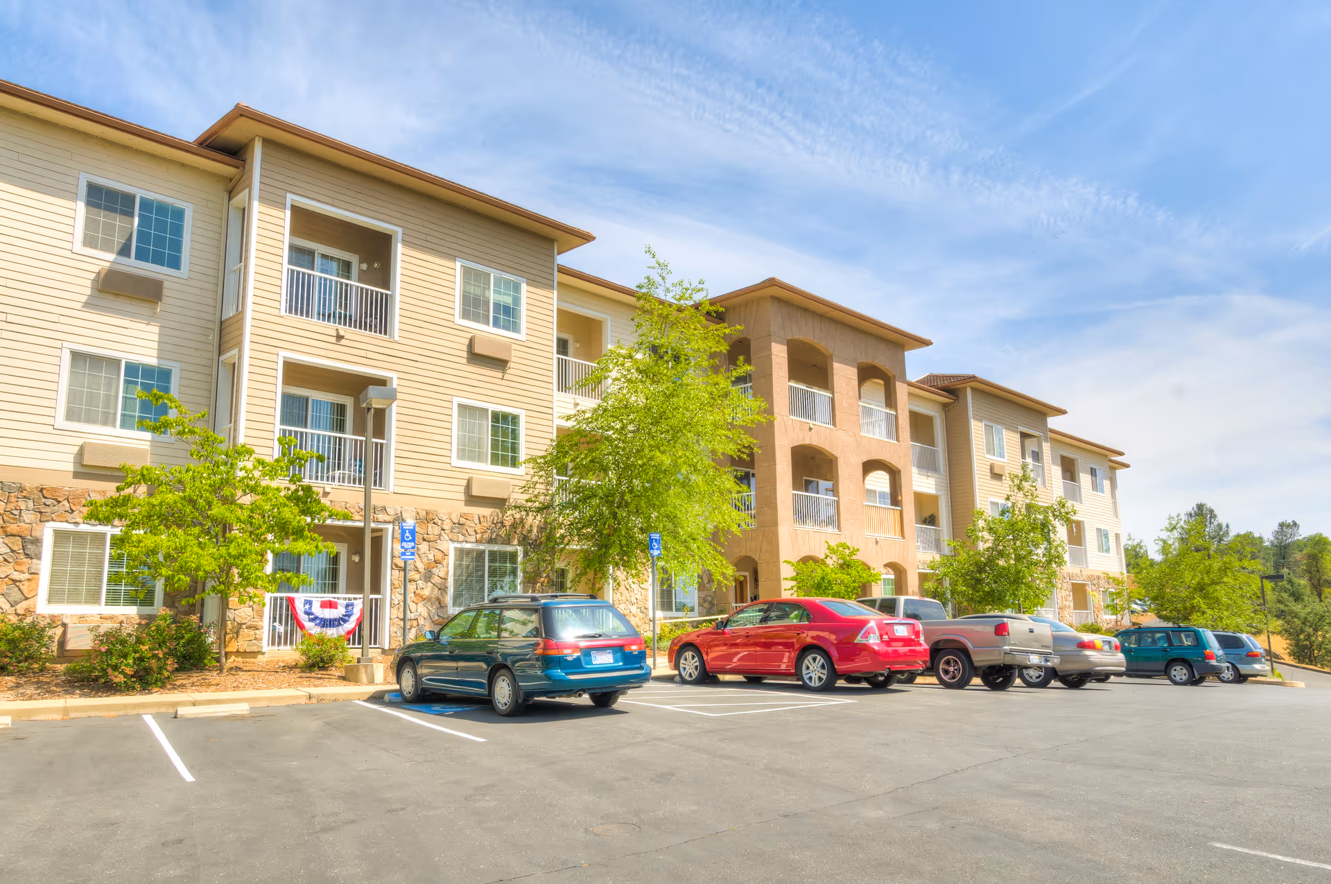 Front exterior of a three-story senior living apartment building with balconies, parked cars, and trees in the parking lot.