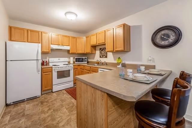 A kitchen with wooden cabinets, a white refrigerator, stove, microwave, and dishwasher. There is a countertop with three place settings and three wooden bar stools. A wall clock is mounted on the wall above the counter.