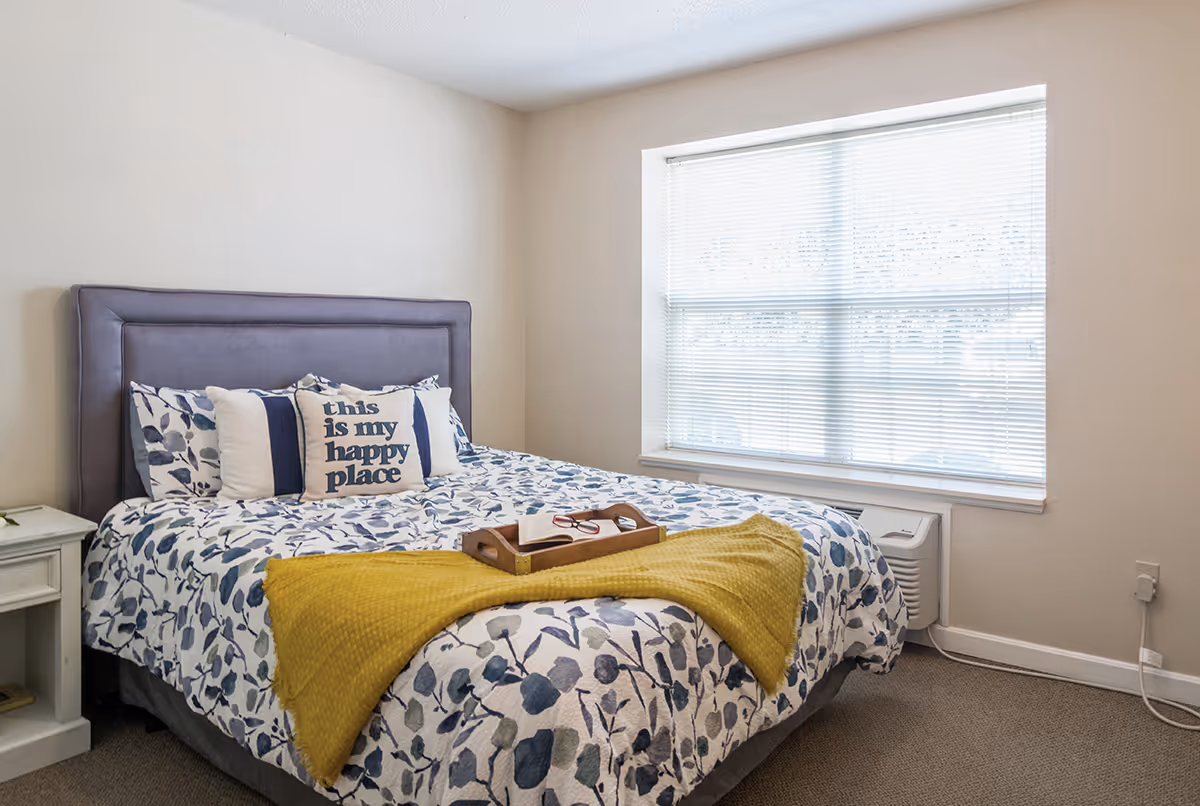 Bright bedroom with a floral-patterned bed, purple headboard, decorative pillows including one that reads "this is my happy place", and a window with blinds.
