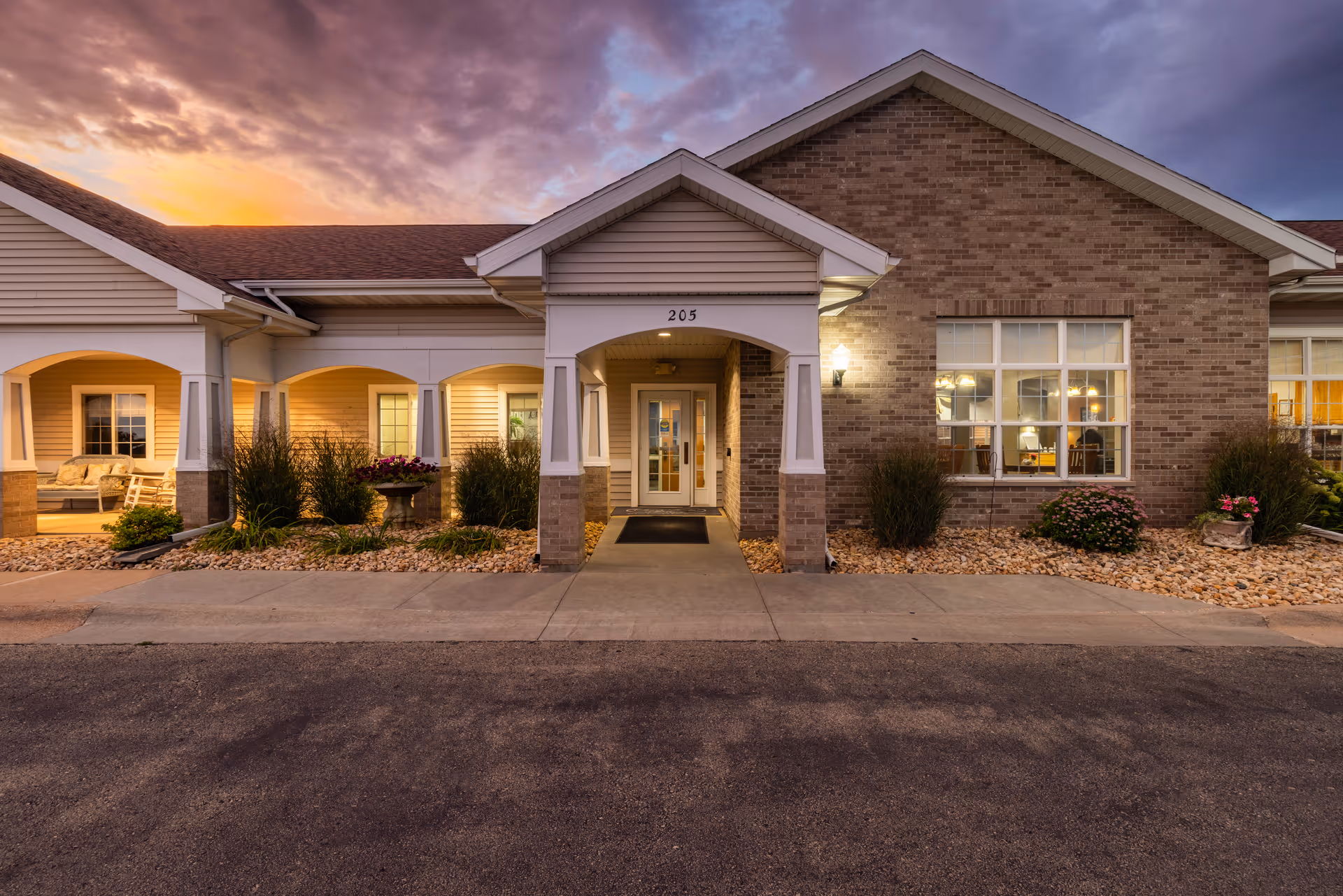 Exterior front view of a single-story senior living facility building at sunset, featuring a covered entrance with the number 205 above it, brick and siding walls, well-maintained landscaping with bushes and flowers, and warm interior lights visible through the windows.