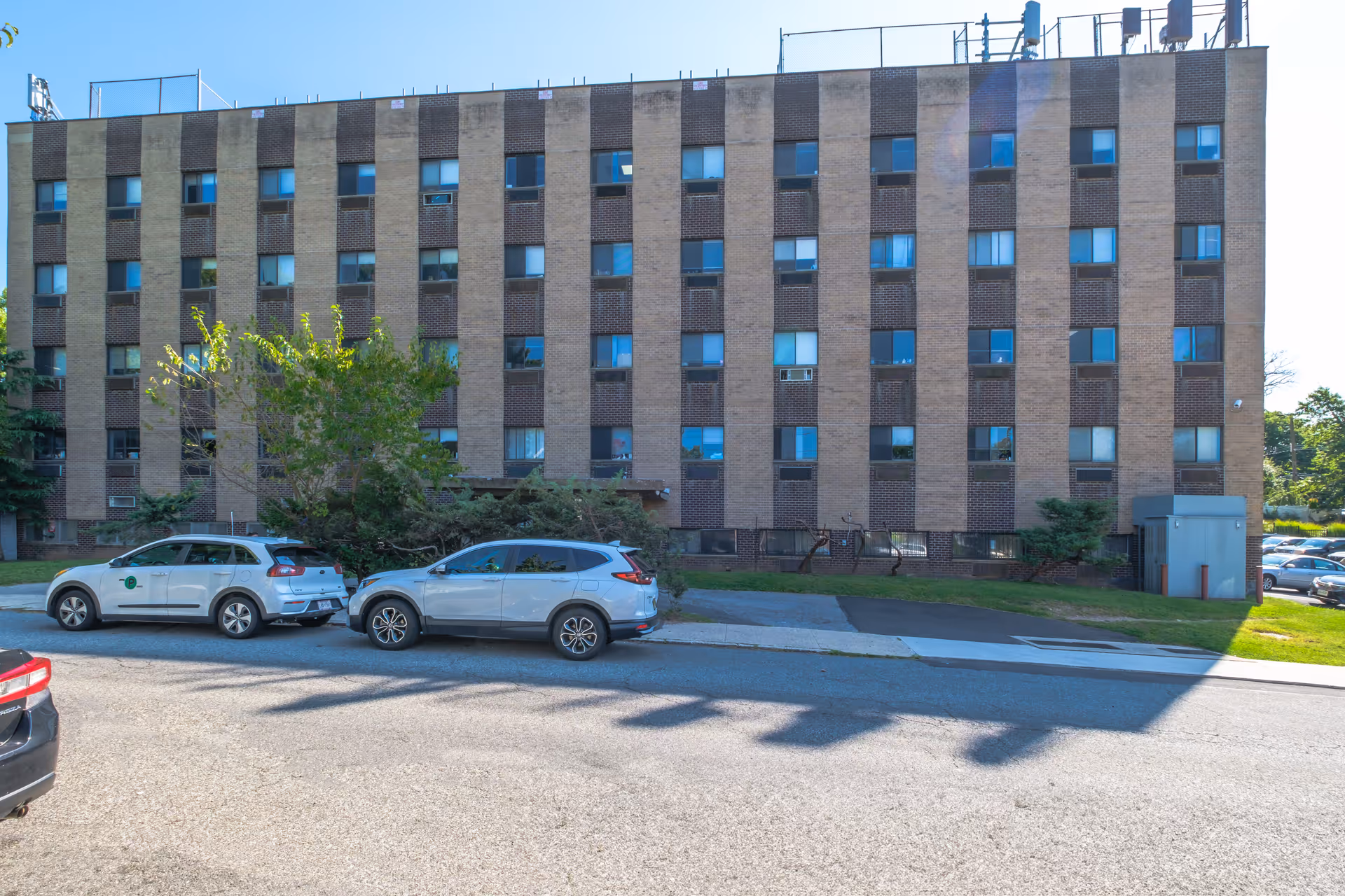 Five-story brick residential building facade with rows of windows, a small lawn, and cars parked along the street.