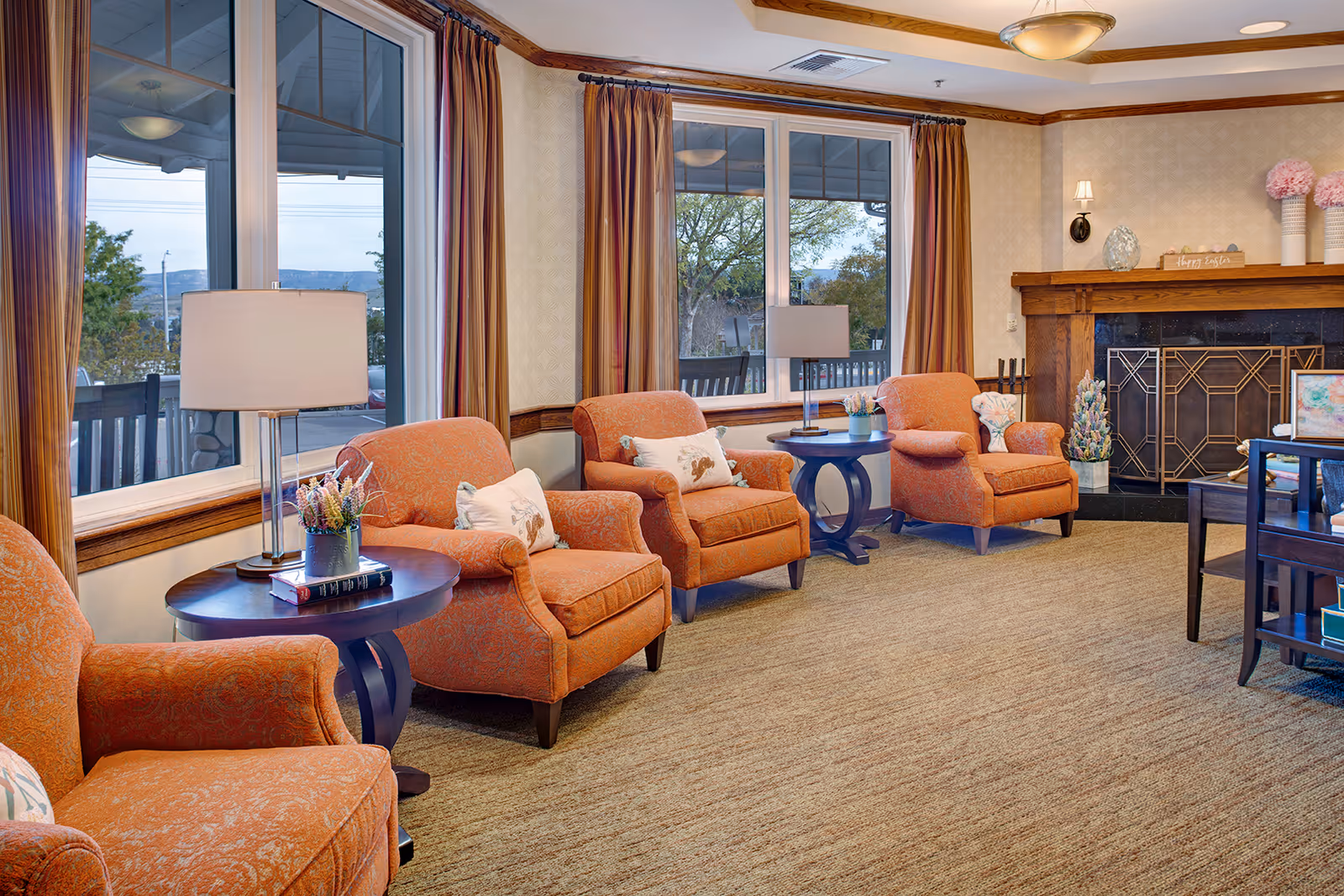 A cozy living room with four orange patterned armchairs arranged around two dark wooden side tables with lamps and decorative items. Large windows with striped curtains let in natural light, and a fireplace with a wooden mantel decorated with flowers and a 'Happy Easter' sign is visible in the background.