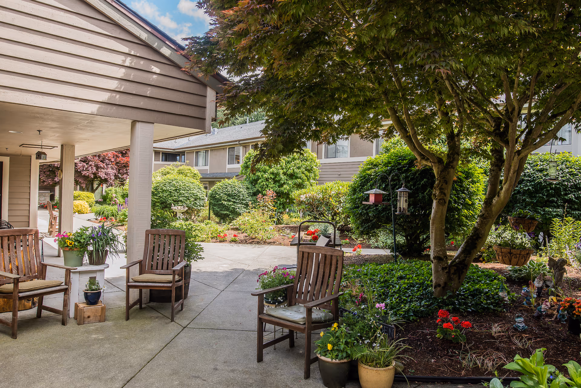 Outdoor seating area with wooden chairs and potted plants under a covered patio next to a garden with trees, bushes, flowers, and bird feeders in front of a residential building.