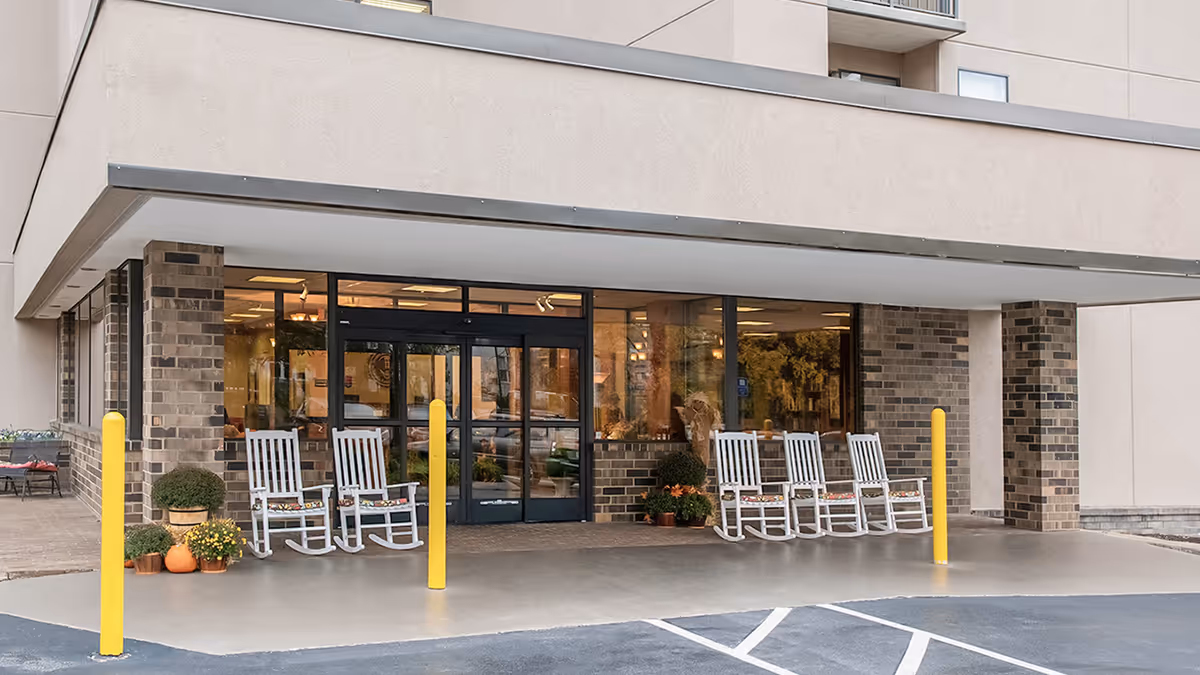 Entrance of a senior living facility with a covered drop-off area. There are six white rocking chairs with cushions arranged in two groups near the glass doors. Several potted plants and flowers are placed near the chairs. The building exterior features beige walls and brick columns. Yellow bollards are positioned near the entrance for protection.