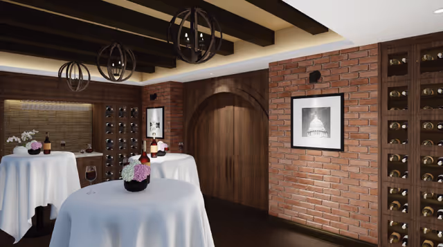 Interior room with brick walls and wooden ceiling beams featuring round tables covered with white tablecloths, each adorned with flower arrangements and wine bottles. Wine racks filled with bottles are built into the walls, and a framed black and white photo hangs on the brick wall.